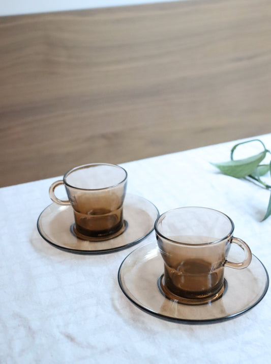 Three brown ceramic tea cups with a sepia tone, placed on matching white saucers, on a white surface with a wooden background.