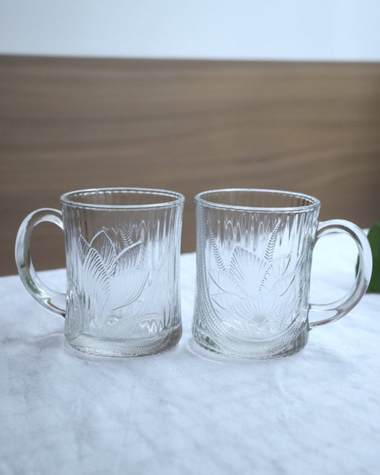 A set of two clear glass coffee mugs with a magnolia flower print, placed on a tablecloth.
