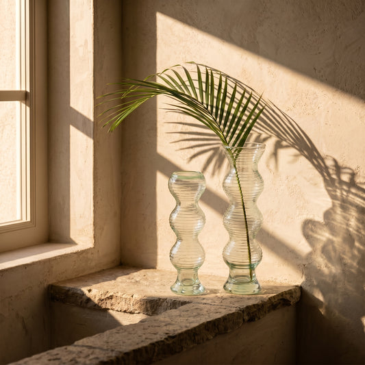 Two glass vases with a plant on a ledge near a window with sunlight casting shadows.