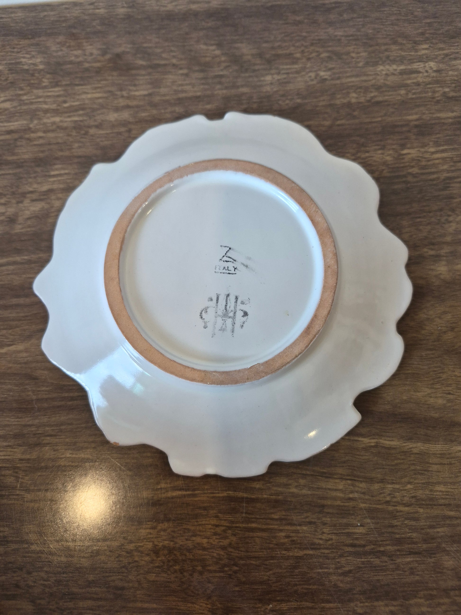 A white ceramic plate with a scalloped rim, photographed from above on a dark brown wooden surface. The inside of the plate shows a hand-written maker's mark and date in black ink.