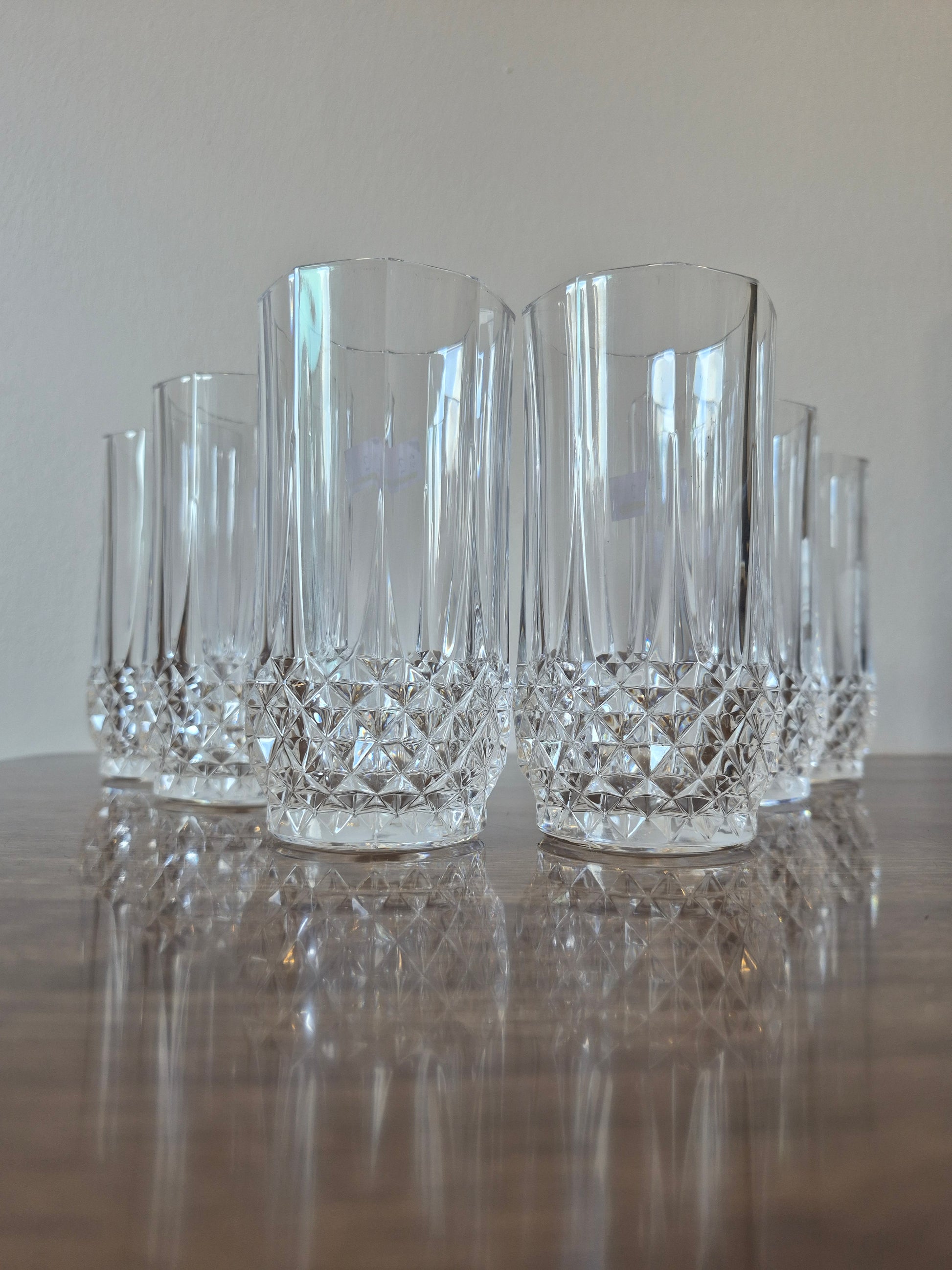 A set of clear cut-crystal tumblers with a faceted diamond-patterned base, arranged on a reflective wooden table against a neutral light gray background.