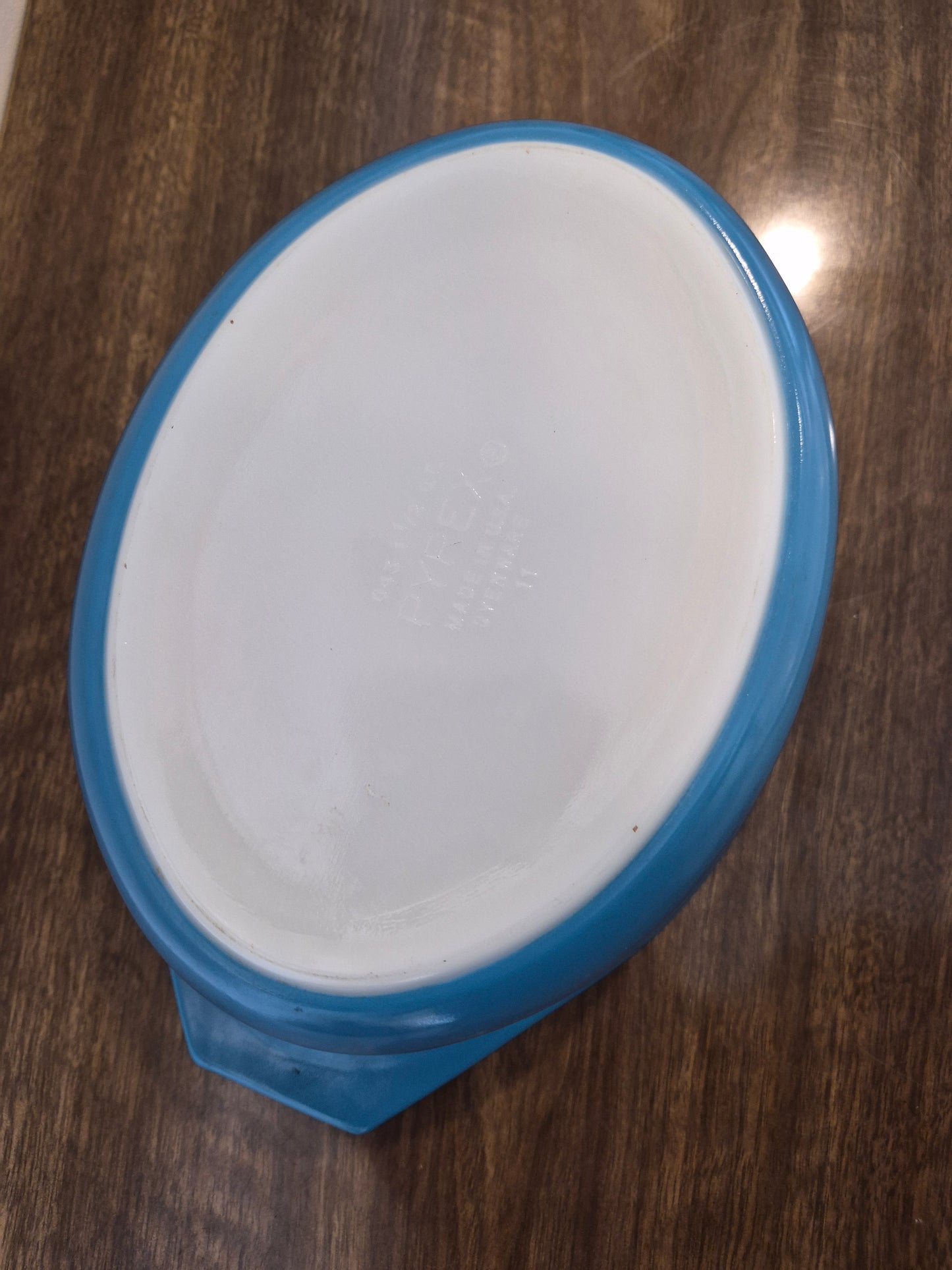 An oval glass baking dish with a blue rim and handle, photographed from above on a dark wood surface.
