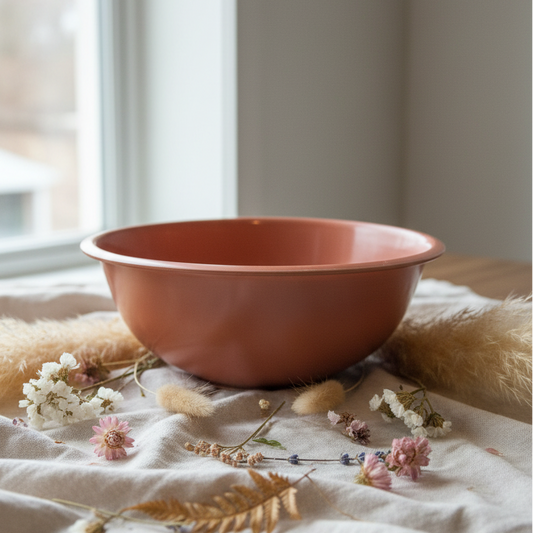 A terracotta-colored ceramic bowl with a wide rim, shown from a side profile against a white wall and a dark wood surface.