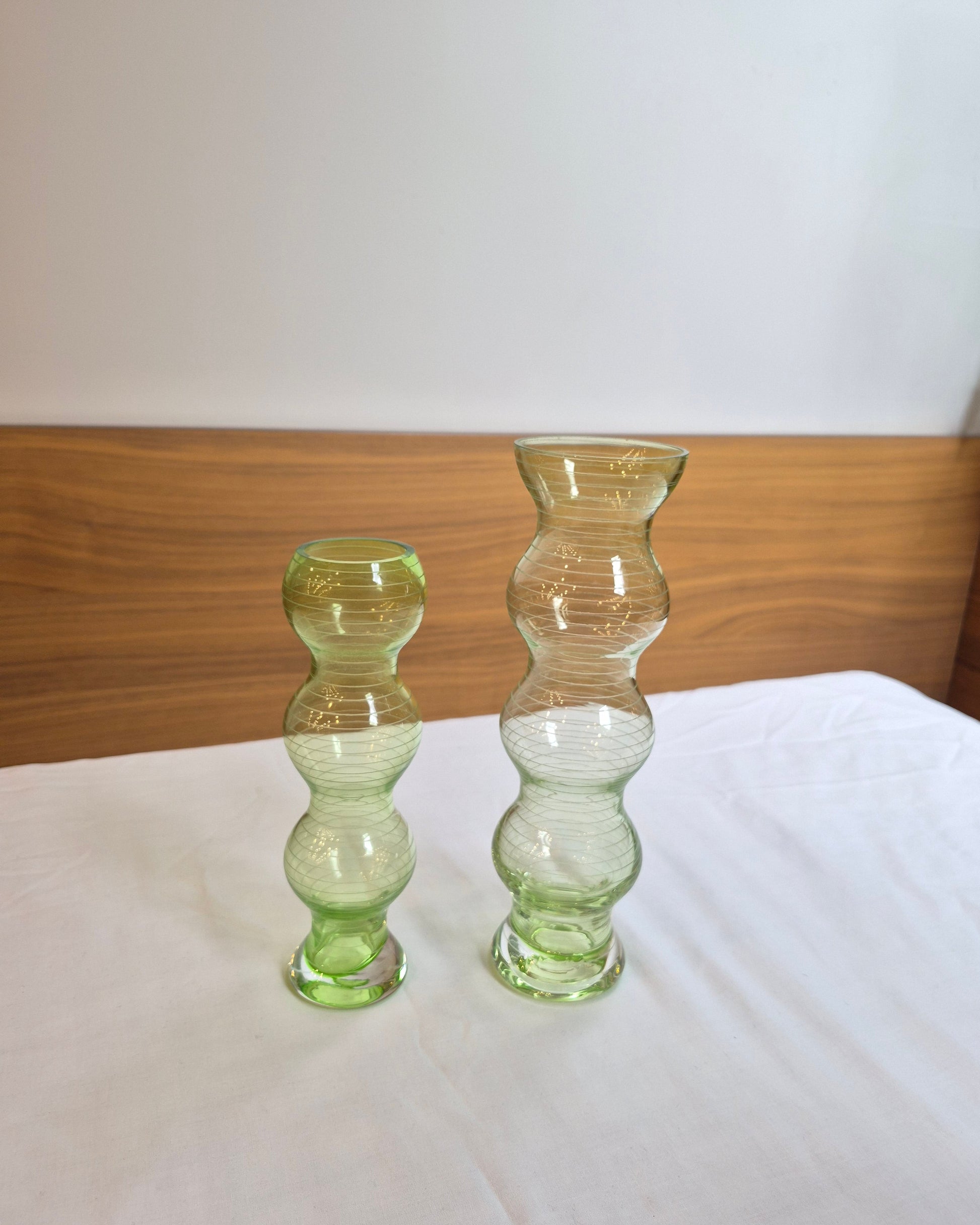 Two green glass vases on a white surface with a wooden background