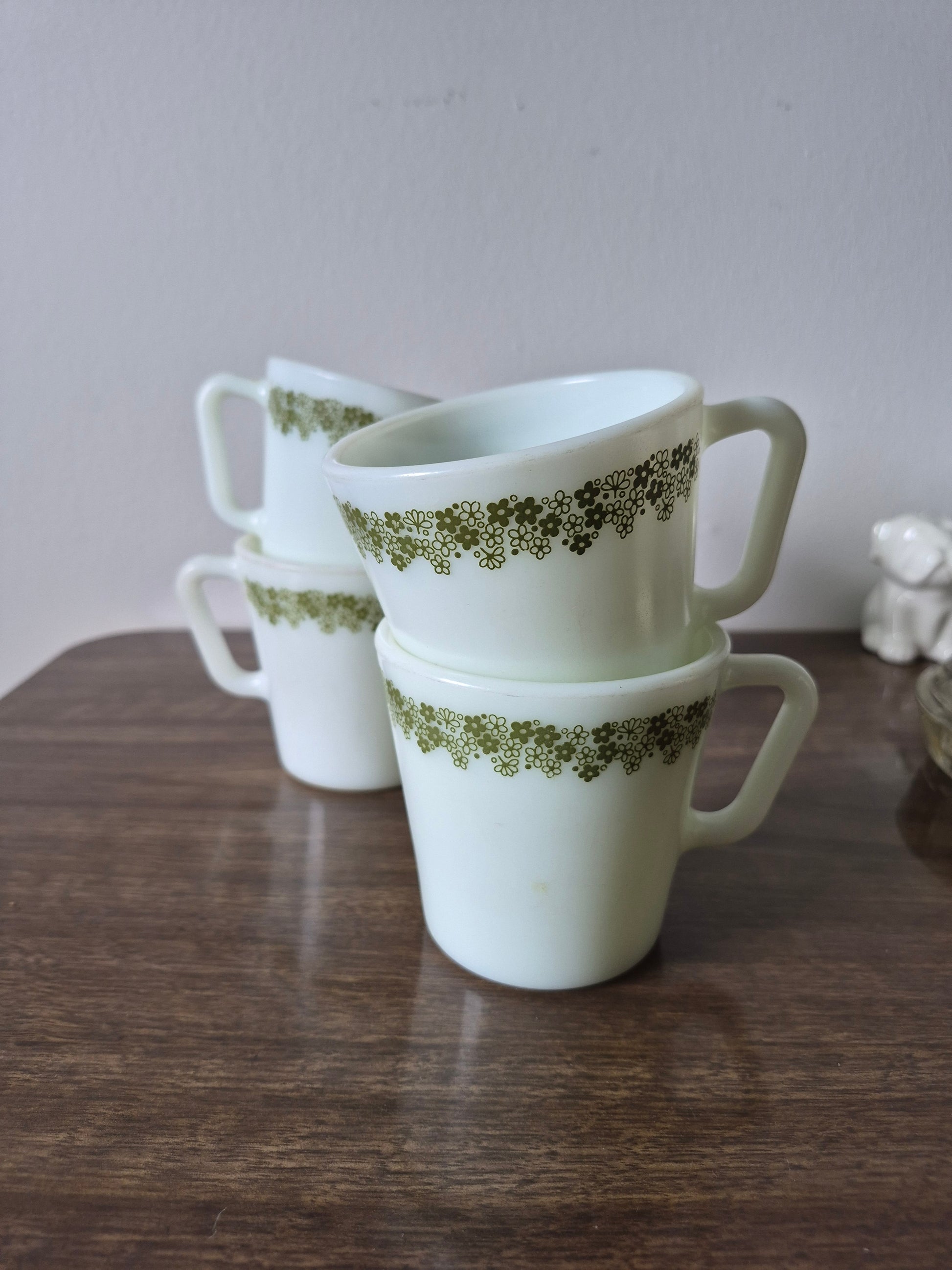 Four white Pyrex mugs with green daisy patterns are stacked on a dark wooden surface against a light background.