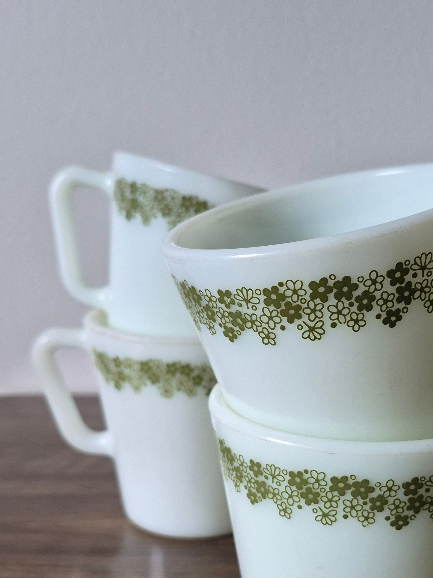 Four white Pyrex mugs with green daisy patterns are stacked on a dark wooden surface against a light background.